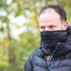 Extremitieshoot_Oct2019-128-scaled-3.jpg Middle-aged man with thinning hair wearing a black face mask and hooded puffer jacket with backpack straps, only his eyes visible as he looks to the right against blurred green-yellow foliage.