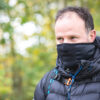 Man outdoors wearing a black neck gaiter pulled up over his mouth and a puffy black jacket with blue-accented backpack straps, looking to the right against a soft-focus green woodland background.