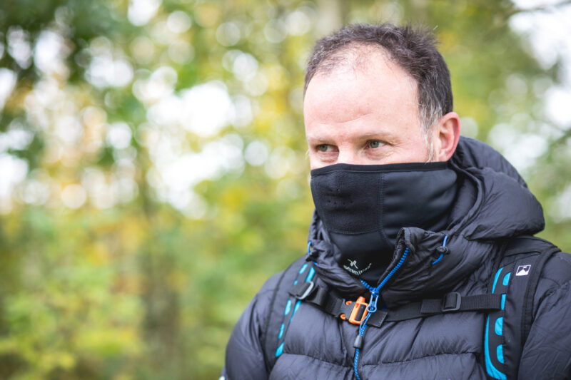 Man outdoors wearing a black neck gaiter pulled up over his mouth and a puffy black jacket with blue-accented backpack straps, looking to the right against a soft-focus green woodland background.