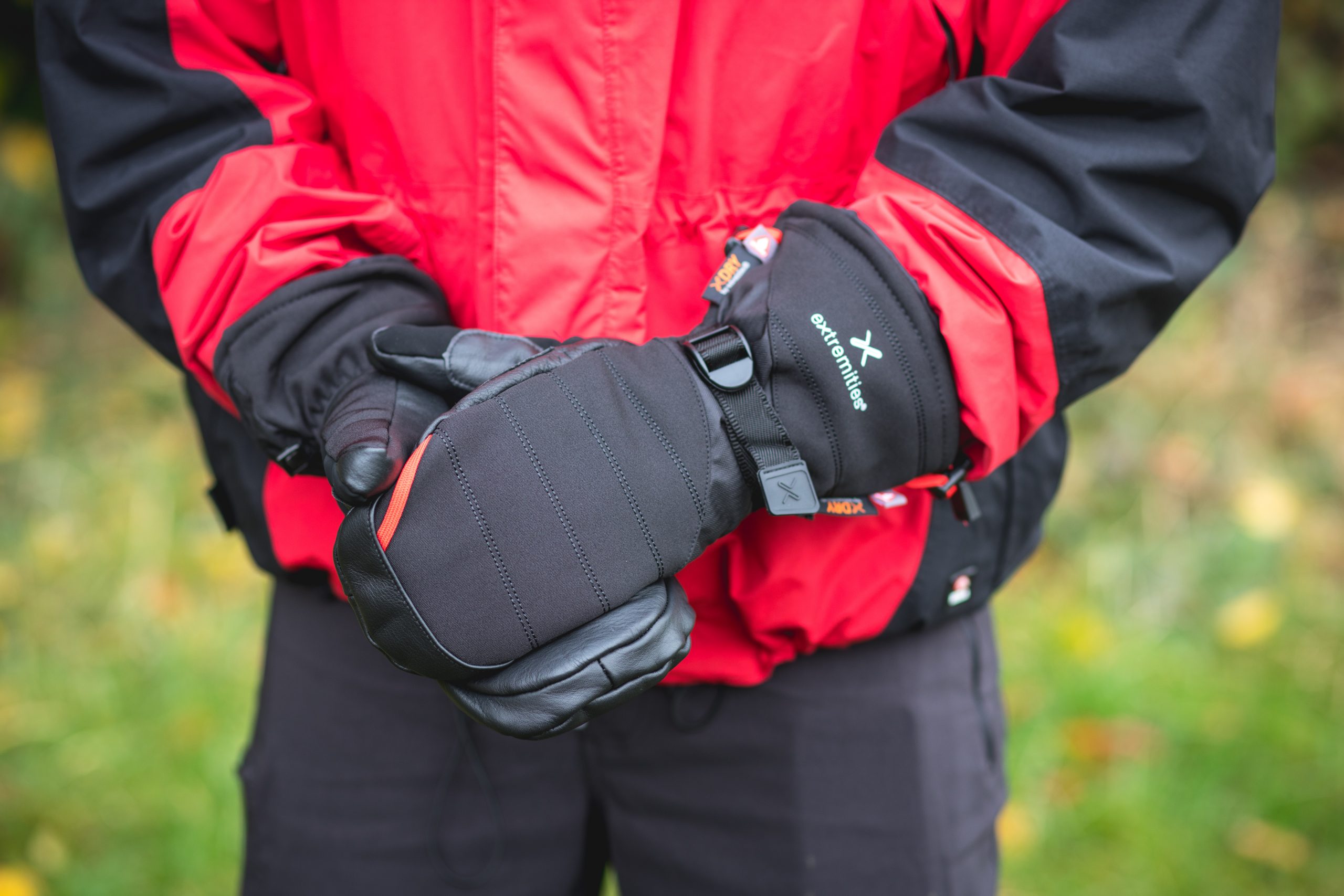 Extremitieshoot_Oct2019-137-scaled-2.jpg Person wearing a red-and-black jacket holding padded black mittens with a strap and visible "extremities" logo against a blurred outdoor background.