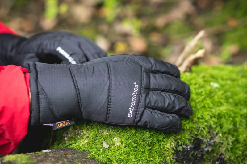 Extremitieshoot_Oct2019-141-scaled-2.jpg Black padded extremities glove lying palm-down on bright green moss with a red jacket cuff visible at the wrist.