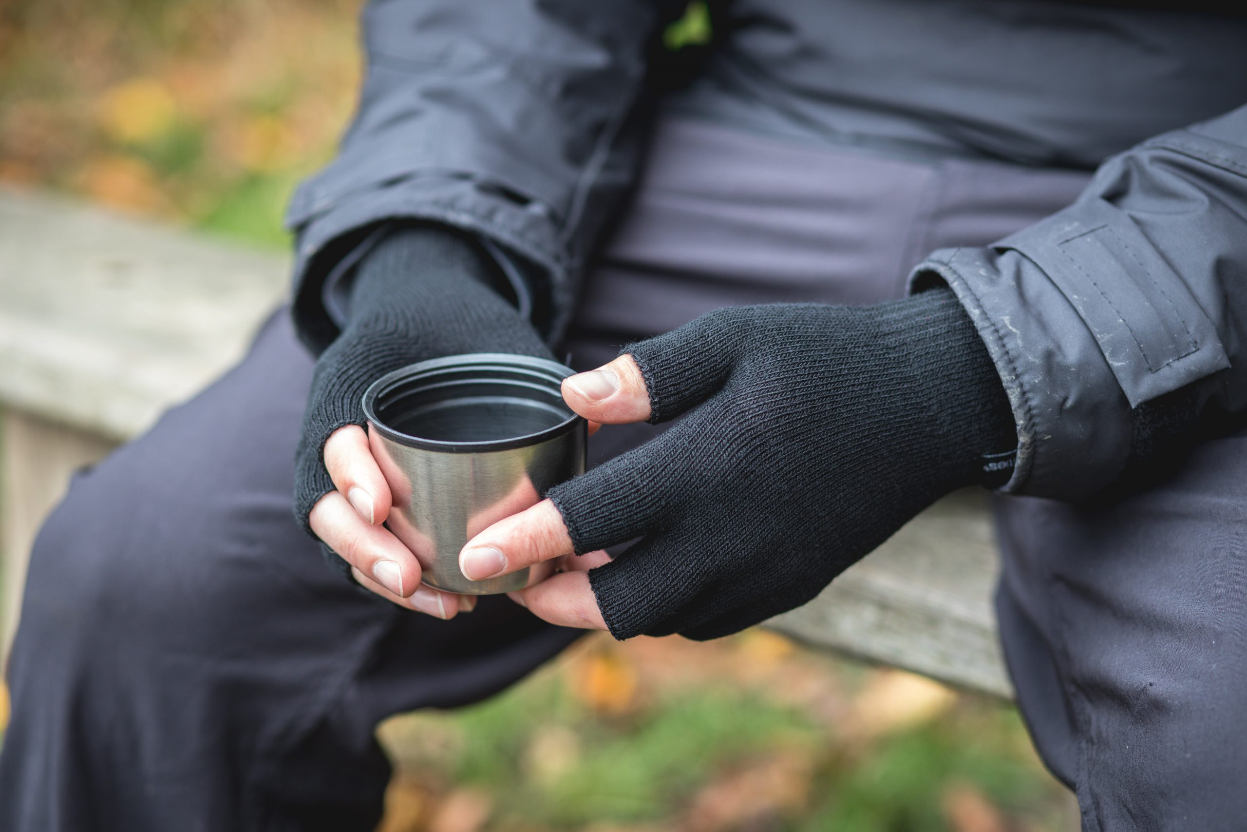 Hands in black fingerless gloves holding a stainless-steel cup while sitting on an outdoor bench, wearing a dark jacket with blurred autumn foliage in the background.