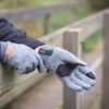 Extremitieshoot_Oct2019-30-1-scaled-2.jpg Hands wearing grey wool gloves with black palm patches and an orange 'XDRY' label resting on a weathered wooden railing, the wearer in a dark jacket on an outdoor boardwalk.