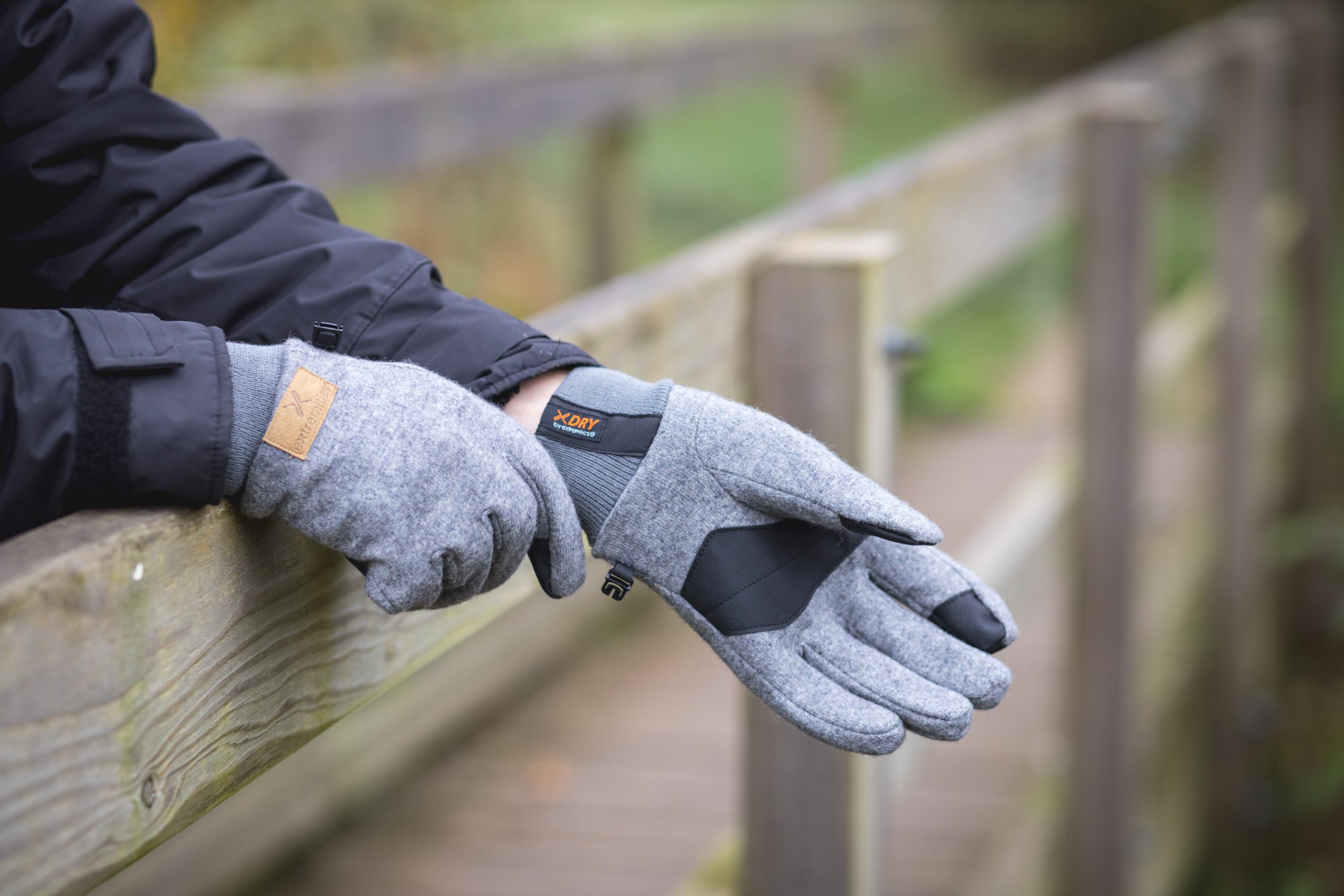 Extremitieshoot_Oct2019-30-1-scaled-2.jpg Hands wearing grey wool gloves with black palm patches and an orange 'XDRY' label resting on a weathered wooden railing, the wearer in a dark jacket on an outdoor boardwalk.