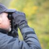 Side-profile of a bearded man in a dark cap and jacket holding binoculars to his eyes with gloved hands against a blurred green woodland background.
