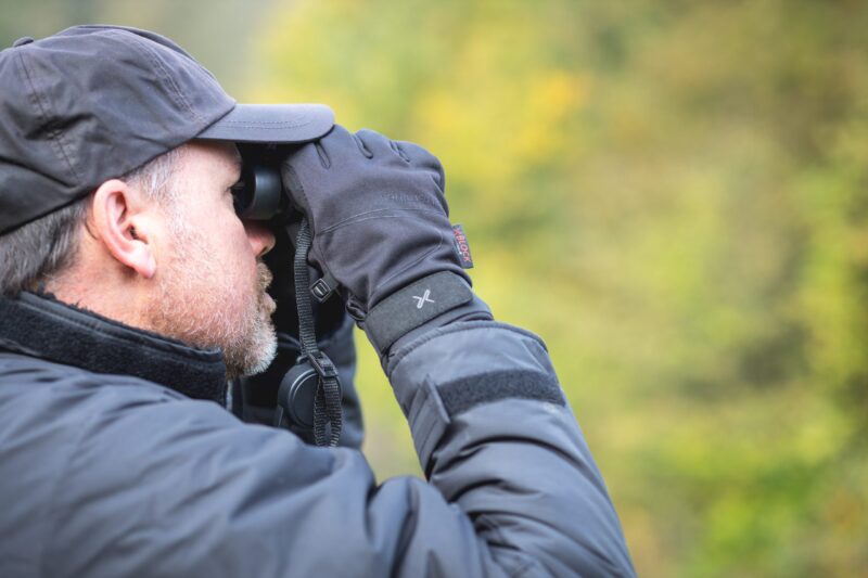 Side-profile of a bearded man in a dark cap and jacket holding binoculars to his eyes with gloved hands against a blurred green woodland background.