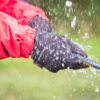 Hands in black "extremities" waterproof gloves and a red jacket sleeve gripping a wet rope as water splashes over them against a blurred green outdoor background.