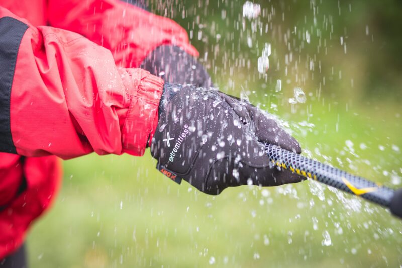 Hands in black "extremities" waterproof gloves and a red jacket sleeve gripping a wet rope as water splashes over them against a blurred green outdoor background.
