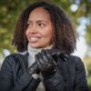 Fernie-Glove-2-scaled-4.jpg Woman with curly hair outdoors, smiling and looking to the left while clasping quilted black leather gloves and wearing a dark jacket over a light turtleneck.