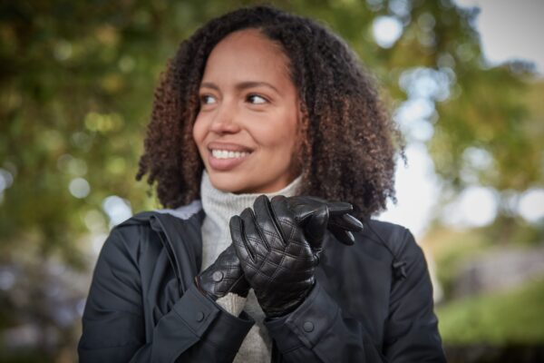 Fernie-Glove-2-scaled-4.jpg Woman with curly hair outdoors, smiling and looking to the left while clasping quilted black leather gloves and wearing a dark jacket over a light turtleneck.