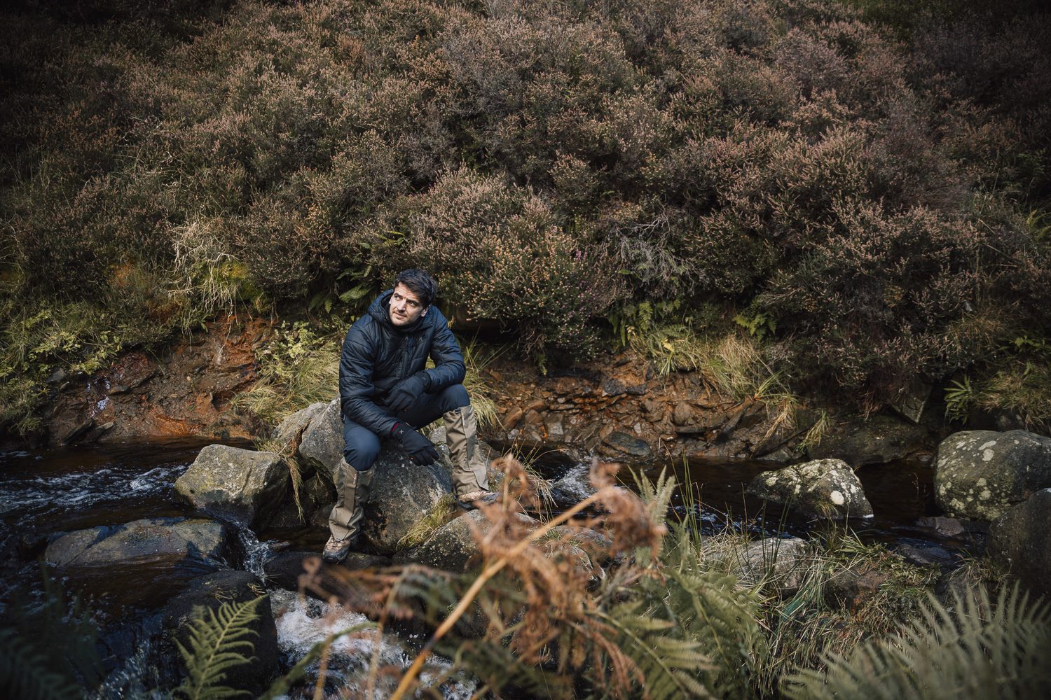 A man in a dark puffer jacket, gloves and gaiters sits on a rock beside a shallow stream, looking upward amid ferns, mossy stones and a dense heather-covered hillside.