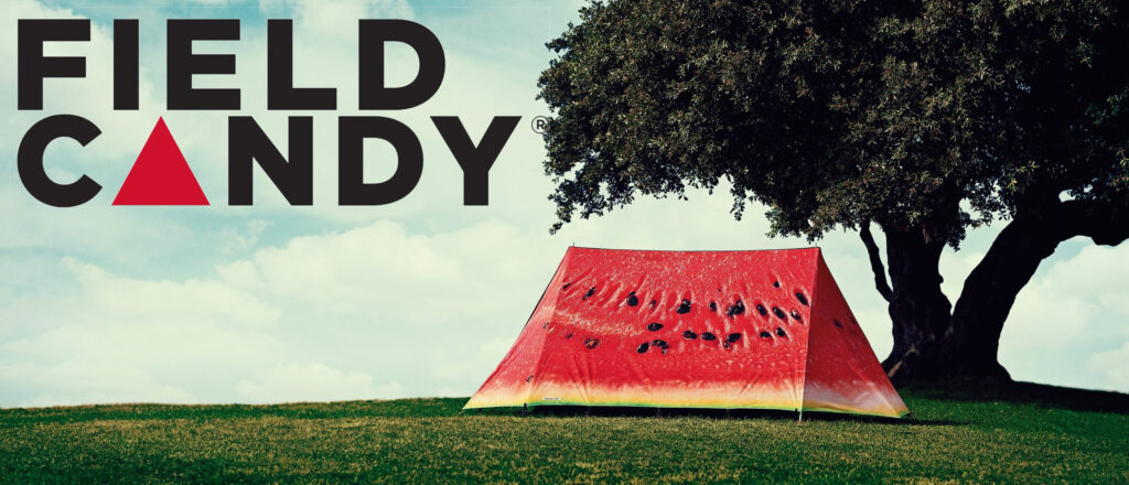 Bright red tent printed like a sliced watermelon with black 'seeds' pitched under a large tree on a grassy hill, with a pale blue sky and a bold FIELD CANDY logo occupying the left side.
