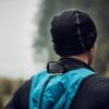 Flux-Beanie-5-scaled-2.jpg Person seen from behind wearing a black beanie and jacket with a bright blue backpack, standing in a misty, blurred forest background.