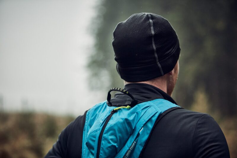 Person seen from behind wearing a black beanie and jacket with a bright blue backpack, standing in a misty, blurred forest background.