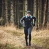 Flux-Beanie-8-scaled-2.jpg A runner wearing black leggings, a black long-sleeve top, gloves and a beanie with a blue hydration backpack, running on a narrow forest path lined with dry grasses and tall conifer trunks.