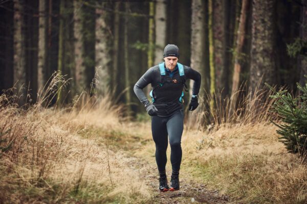 Flux-Beanie-8-scaled-2.jpg A runner wearing black leggings, a black long-sleeve top, gloves and a beanie with a blue hydration backpack, running on a narrow forest path lined with dry grasses and tall conifer trunks.