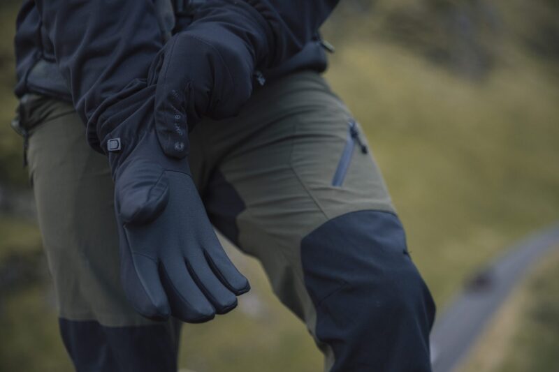 Focus-Glove-2-3.jpg Close-up of a person wearing black insulated gloves and olive-green outdoor trousers with reinforced black knee panels and a zipped thigh pocket, hands extended against a blurred grassy hillside background.