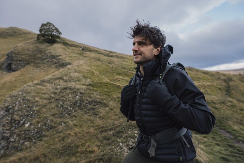 Focus-Glove-5-3.jpg A man in a black puffer jacket and gloves carrying a backpack hikes across grassy rolling hills under an overcast sky with a solitary tree on a distant ridge.