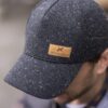 Furnace-Cap-2-3.jpg Dark grey flecked baseball cap with a tan leather 'extremities' patch, worn tilted forward on a man's head with his face blurred in the background.