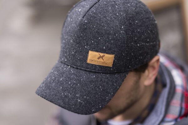 Furnace-Cap-2-3.jpg Dark grey flecked baseball cap with a tan leather 'extremities' patch, worn tilted forward on a man's head with his face blurred in the background.