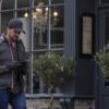 Furnace-Cap-and-Bora-Contact-Gloves-4-3.jpg Man in a dark cap, padded vest, checked shirt and gloves walking past a café window while looking at his smartphone, a chandelier visible inside and potted trees by the entrance
