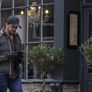 Man in a dark cap, padded vest, checked shirt and gloves walking past a café window while looking at his smartphone, a chandelier visible inside and potted trees by the entrance