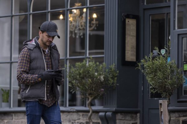 Furnace-Cap-and-Bora-Contact-Gloves-4-3.jpg Man in a dark cap, padded vest, checked shirt and gloves walking past a café window while looking at his smartphone, a chandelier visible inside and potted trees by the entrance