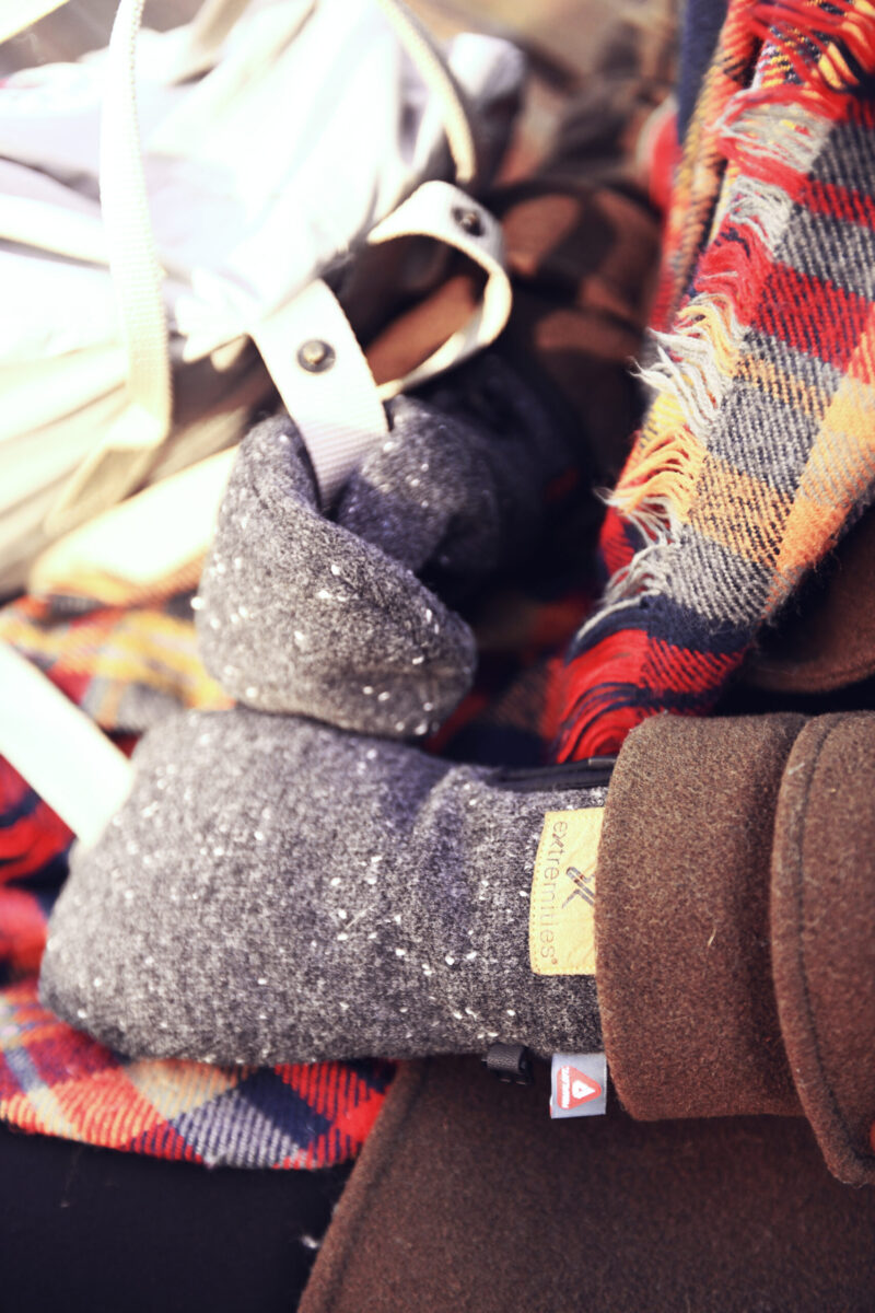 Furnace Mitt Cozy winter accessories featuring a close-up of a hand wearing a gray knitted glove, surrounded by a colorful plaid scarf and a brown jacket, perfect for cold weather fashion.