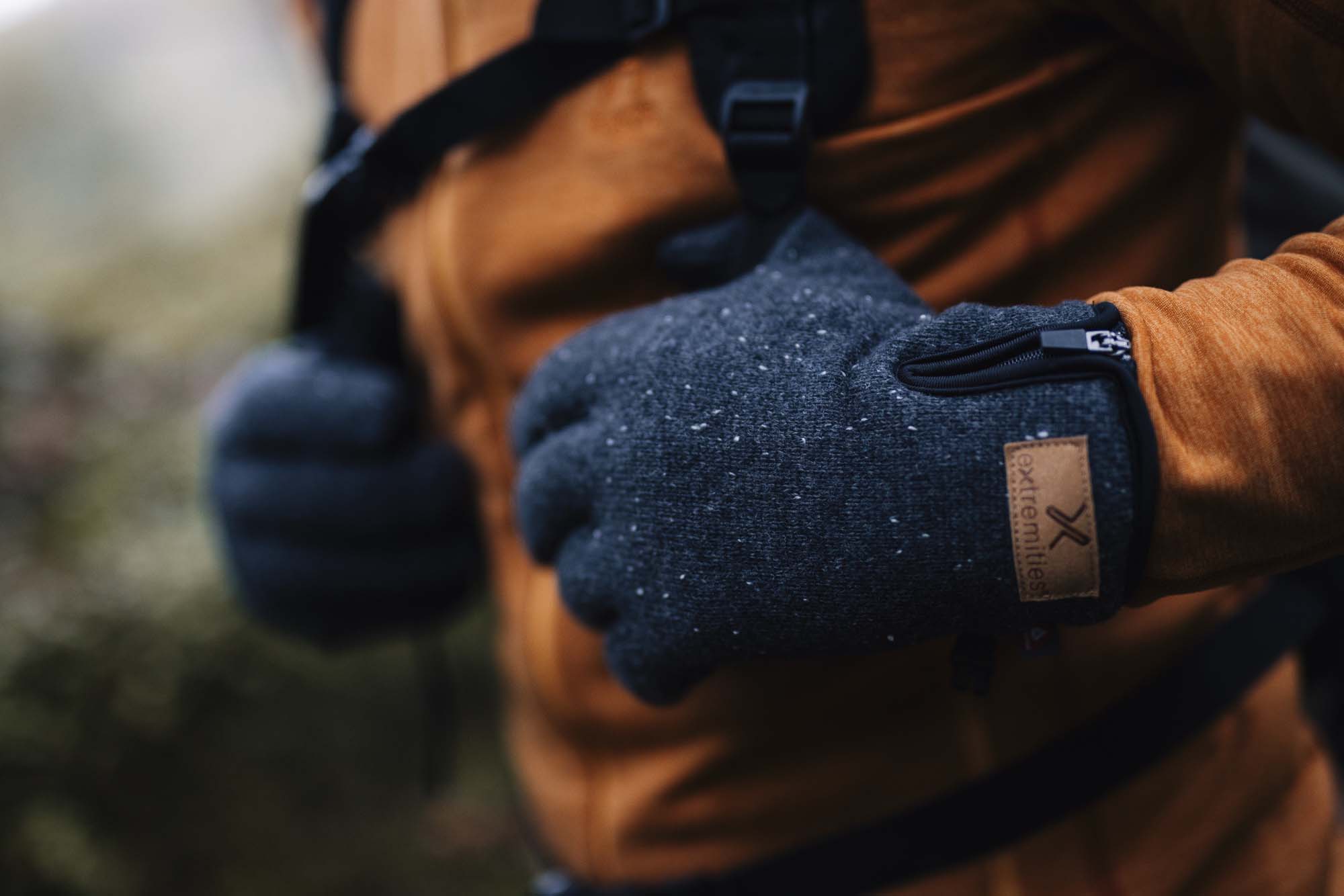Close-up of hands in speckled dark grey gloves with a small zipped pocket and leather logo patch, gripping backpack straps over an orange outdoor jacket against a blurred woodland background.