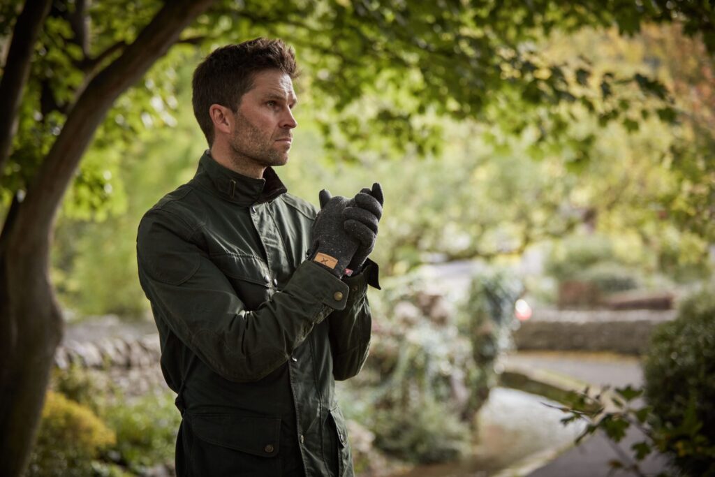 Man in a dark green jacket and grey gloves stands under leafy trees, clasping his hands and looking pensively to the left with a blurred garden path and greenery behind him.