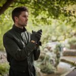 Man in a dark green jacket and grey gloves stands under leafy trees, clasping his hands and looking pensively to the left with a blurred garden path and greenery behind him.