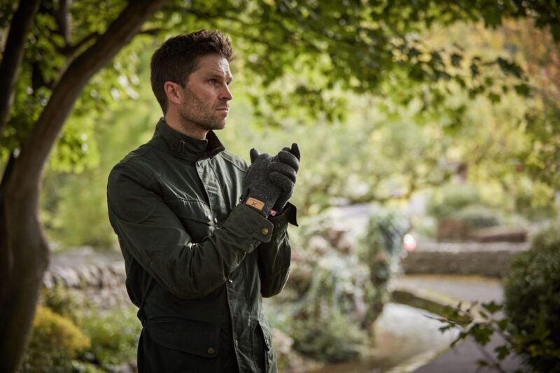Furnace-Pro-Gloves-2-scaled-4.jpg Man in a dark green jacket and grey gloves stands under leafy trees, clasping his hands and looking pensively to the left with a blurred garden path and greenery behind him.