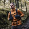Man in an orange fleece jacket and grey speckled gloves adjusting his backpack's chest strap while walking on a moss-covered woodland path with trees and autumnal foliage.