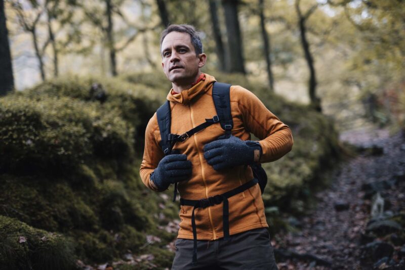 Furnace-Pro-Gloves-24-3.jpg Man in an orange fleece jacket and grey speckled gloves adjusting his backpack's chest strap while walking on a moss-covered woodland path with trees and autumnal foliage.