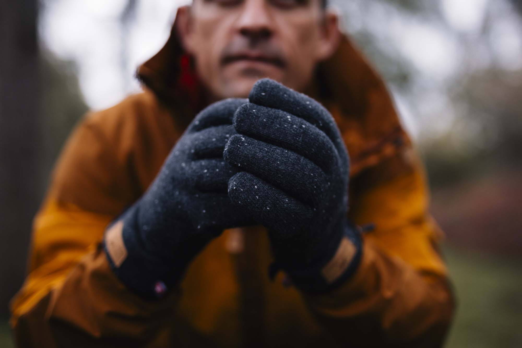 Close-up of a person in an orange jacket holding their dark flecked winter gloves together, with their face blurred in the background.