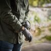 Torso of a person in a dark green jacket and blue jeans adjusting grey wool gloves while standing on a blurred riverside path.