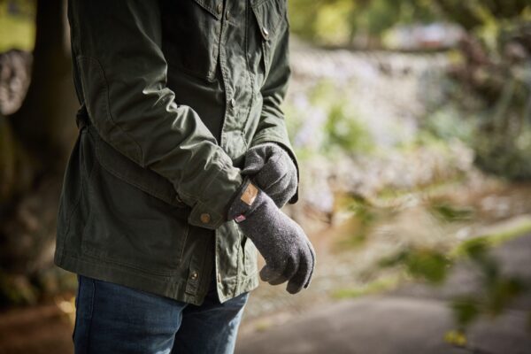Torso of a person in a dark green jacket and blue jeans adjusting grey wool gloves while standing on a blurred riverside path.