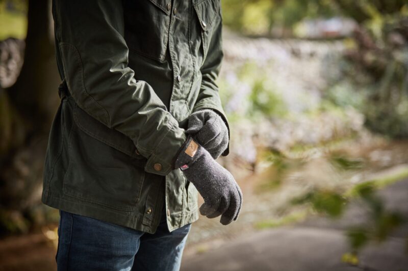 Furnace-Pro-Gloves-scaled-4.jpg Torso of a person in a dark green jacket and blue jeans adjusting grey wool gloves while standing on a blurred riverside path.
