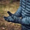 A person in a wet dark blue quilted puffer jacket and black gloves, both dotted with raindrops, holds out cupped hands against a blurred woodland background.