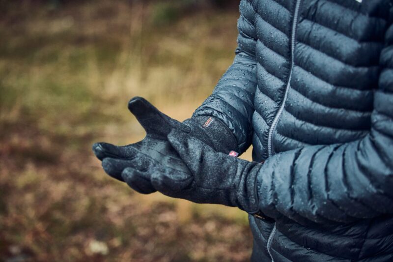 Furnace-Ultra-Charcoal-Lifestyle-2-scaled-4.jpg A person in a wet dark blue quilted puffer jacket and black gloves, both dotted with raindrops, holds out cupped hands against a blurred woodland background.