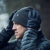 Profile of a man outdoors in light rain adjusting a dark ribbed beanie with a small white X logo while wearing black gloves and a wet quilted puffer jacket against a blurred forest background.