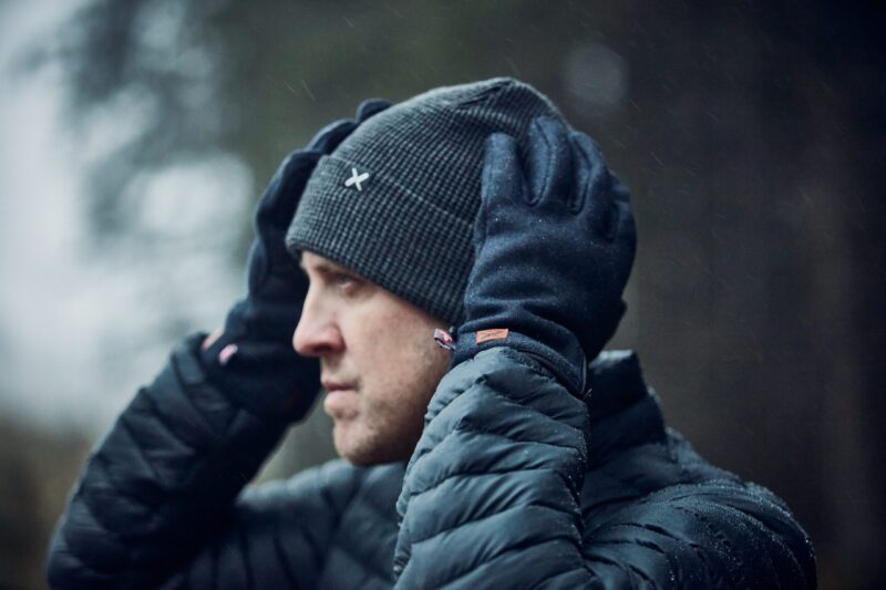 Furnace-Ultra-Navy-Lifestyle-2-scaled-4.jpg Profile of a man outdoors in light rain adjusting a dark ribbed beanie with a small white X logo while wearing black gloves and a wet quilted puffer jacket against a blurred forest background.