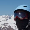 Close-up of a person wearing a light-blue ski helmet, red reflective goggles and a black face mask, with snow-covered mountain peaks behind and skiers reflected in the goggles.