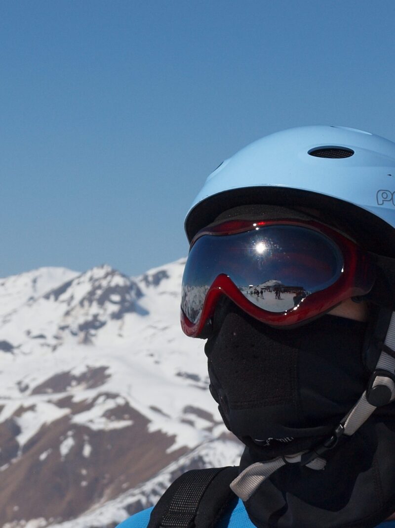Close-up of a person wearing a light-blue ski helmet, red reflective goggles and a black face mask, with snow-covered mountain peaks behind and skiers reflected in the goggles.