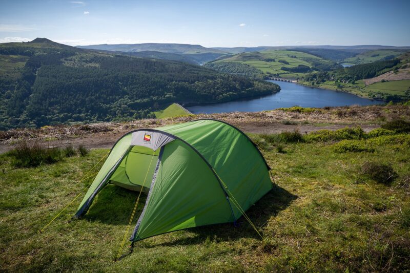 Small green tent pitched on a grassy hillside overlooking a reservoir with a dam and arched bridge, surrounded by wooded slopes and rolling green fields under a clear blue sky.