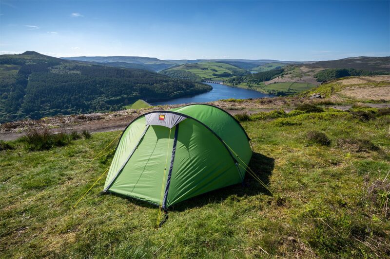 Green dome tent pitched on a grassy hillside overlooking a blue reservoir and rolling patchwork hills under a clear blue sky.