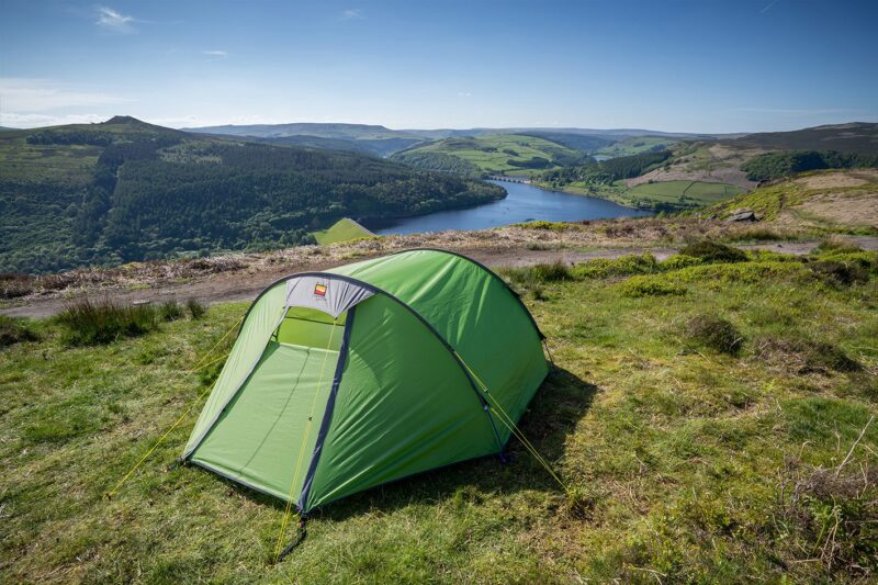 Bright green dome tent pitched on a grassy hillside overlooking a winding reservoir, wooded valleys and rolling green hills beneath a clear blue sky.