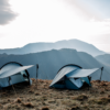 Halny-Elite-1-LS-2-3.webp Two lightweight blue camping tents pitched on a windswept grassy ridge with trekking poles propping their rainflies, set against layered mountain silhouettes under a pale sky.