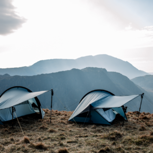 Two lightweight blue camping tents pitched on a windswept grassy ridge with trekking poles propping their rainflies, set against layered mountain silhouettes under a pale sky.
