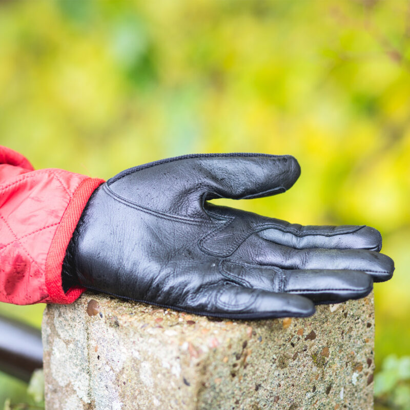 Black leather glove resting palm-up on a rough concrete post, a red quilted jacket cuff visible at the wrist and a soft green-yellow blurred background.